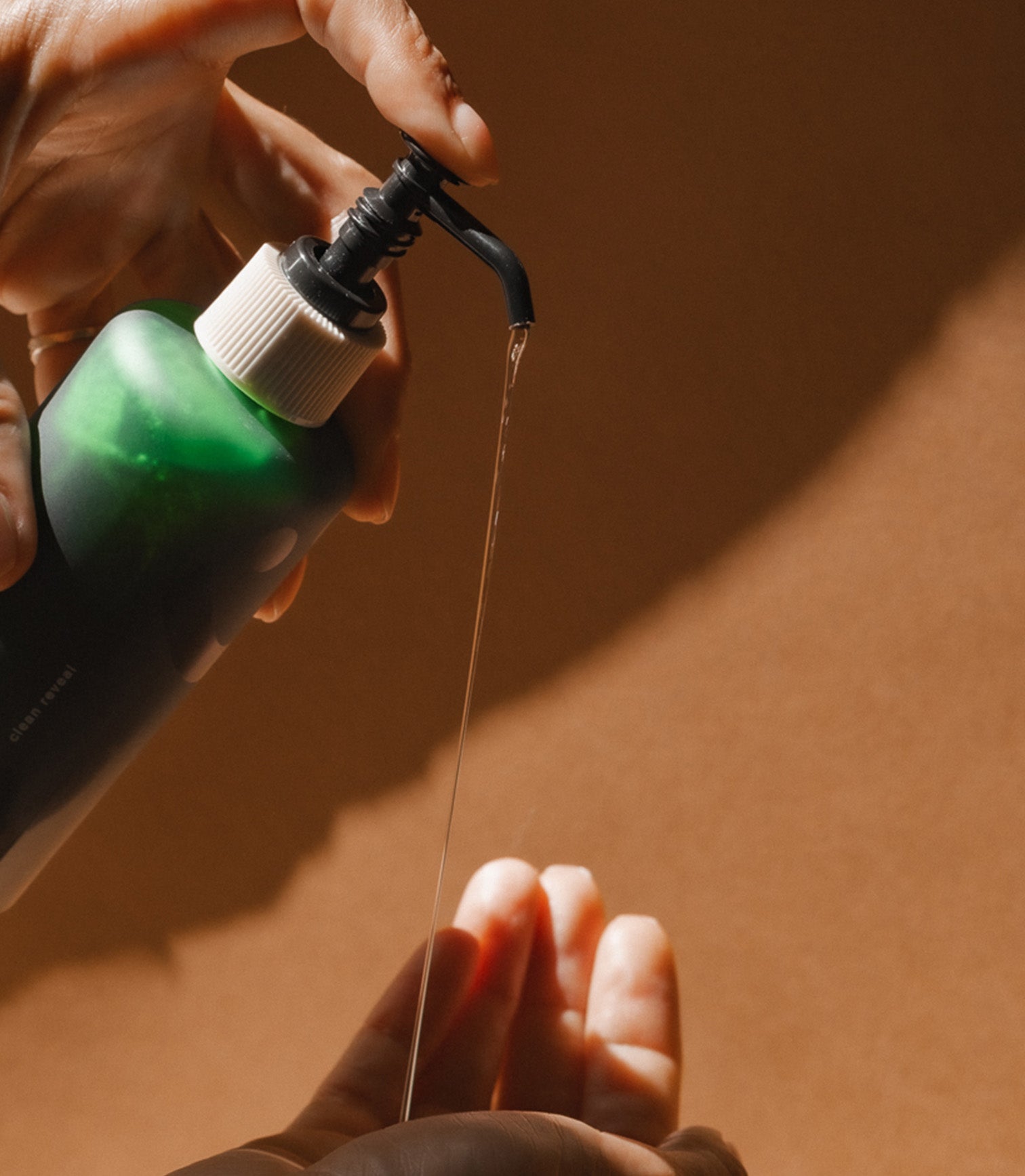 Person holding a green bottle with a pump, dispensing liquid onto another hand against a brown background